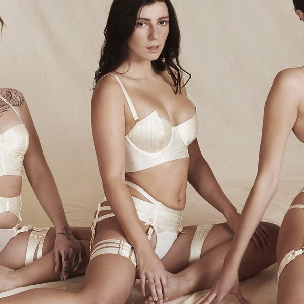 Webbed cream suspender belt by Bordelle. This product is made of elastic and small satin bows with 24 carat gold plated clasps and adjustments. The product is made up of six elastics that wrap around the hips. Two satin elastics are placed vertically with two small bows at the end that suspend the garter belt. At the back a knot is placed on the top of the string of the thong with a golden ring where all the elastics join.
