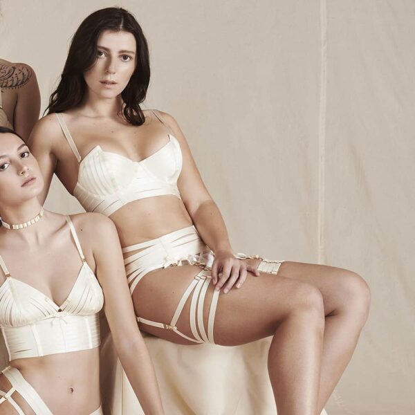 Webbed cream suspender belt by Bordelle. This product is made of elastic and small satin bows with 24 carat gold plated clasps and adjustments. The product is made up of six elastics that wrap around the hips. Two satin elastics are placed vertically with two small bows at the end that suspend the garter belt. At the back a knot is placed on the top of the string of the thong with a golden ring where all the elastics join.