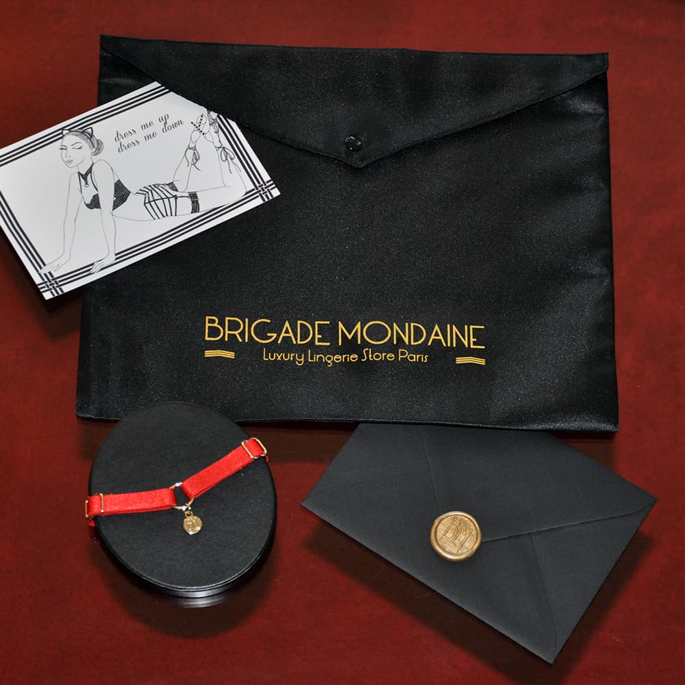 Ici vous pouvez voir le Pack cadeau luxe de la marque Brigade Mondaine. Dedans il y a un chocker rouge avec sa pochette et une carte signée et dédicacée juste pour vous. Tout cela est contenu dans une pochette en soie noire.