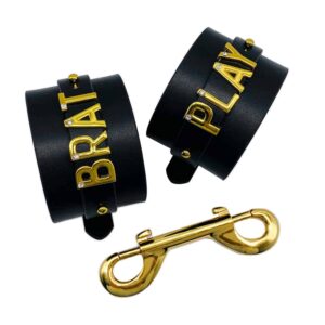 Pair of handcuffs for wrists personalized with writing BRAT PLAY on a black Italian leather with letters and hangers in 24 carat gold plated and small stones on each of the letters from the collaboration UPKO X Brigade Mondaine presented on a white background at Brigade Mondaine