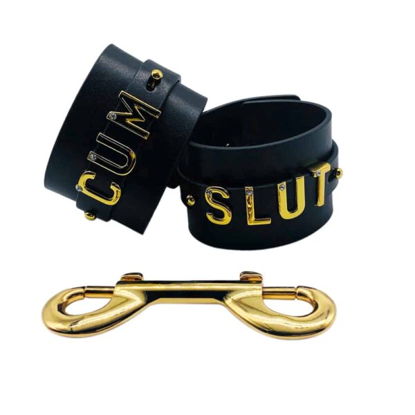 Pair of handcuffs for wrists personalized with writing CUM SLUT on a black Italian leather with letters and hangers in 24 carat gold plated and small stones on each of the letters from the collaboration UPKO X Brigade Mondaine presented on a white background at Brigade Mondaine