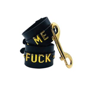 Pair of handcuffs for wrists personalized with writing FUCK ME on a black Italian leather with letters and hangers in 24 carat gold plated and small stones on each of the letters from the collaboration UPKO X Brigade Mondaine presented on a white background at Brigade Mondaine