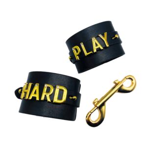 Пара наручников для запястий, персонализированных с надписью PLAY HARD на черной итальянской коже с буквами и вешалками из 24-каратного золота и мелкими камнями на каждом из букв сотрудничества UPKO X Brigade Mondaine, представленных на белом фоне на Brigade Mondaine.