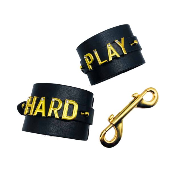 Pair of handcuffs for wrists personalized with writing PLAY HARD on a black Italian leather with letters and hangers in 24 carat gold plated and small stones on each of the letters from the collaboration UPKO X Brigade Mondaine presented on a white background at Brigade Mondaine