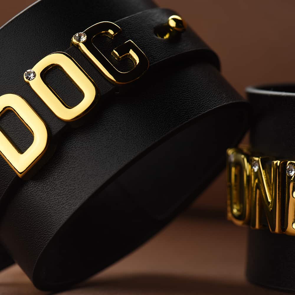 Handcuffs for wrists in black Italian leather seen in close-up showing the word DOG with 24 carat gold-plated letters with stones inlaid in each letter used as an idea for the collaboration UPKO X Brigade Mondaine