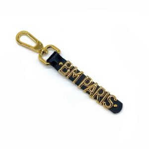 Gold and leather accessory Keyring BM Paris in limited edition collaboration between UPKO and Brigade Mondaine in leather with gold lettering with a small shiny diamond all on a white background