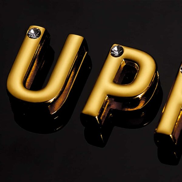 Die Buchstaben U und P aus 24 Karat vergoldetem Gold mit Intarsien aus kleinen glänzenden Steinen auf einem schwarzen Hintergrund aus der Nähe betrachtet für die UPKO X Brigade Mondaine Kollaboration bei Brigade Mondaine