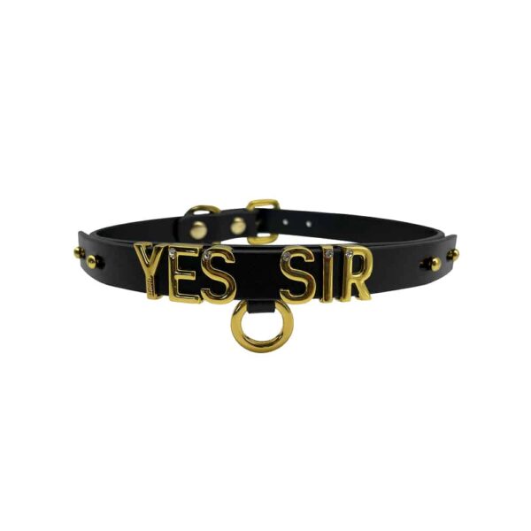 Collier / choker en cuir noir italien souple avec accroche et lettres en or plaqué 24 carats et une petite pierre incrustée sur chacune des lettres écrivant le mot YES SIR de la collection UPKO X Brigade Mondaine présenté sur fond Blanc disponible chez Brigade Mondaine