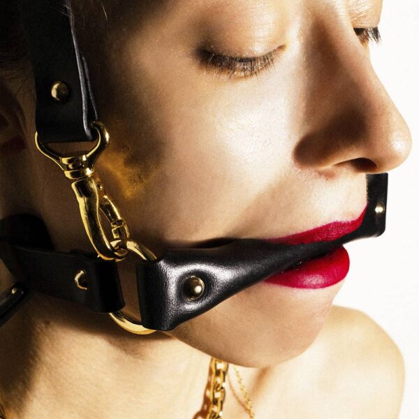 Gag head harness in black leather from Elif Domanic. A strap placed at the level of the mouth is maintained by two golden rings to another strap which is placed at the back of the skull. Another strap much longer is attached to the whole by a loop and loops.