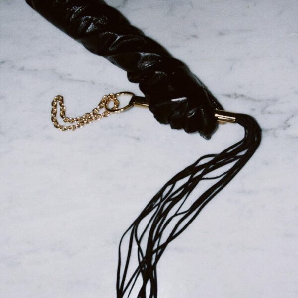 Cybele whip from the brand Elif Domanic. This product is made of black leather and golden brass. The handle is made of golden brass with a ring at the end where a chain made of an accumulation of golden balls is held.