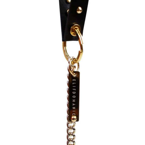 Venus key ring from Elif Domanic. A black leather strap decorated with golden studs is attached to a golden brass ring where a silver metal leash with a snap hook at its end is also present. A golden plate is embossed with the inscriptions ELIF DOMANIC is also present on the ring.