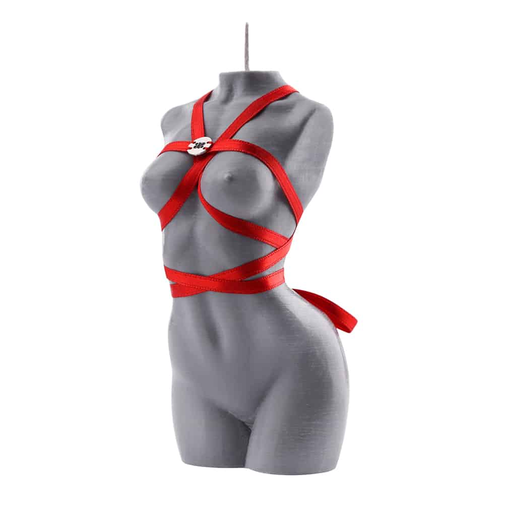 Vela de la marca Baed Stories. Esta vela gris representa el cuerpo de una mujer desnuda arqueada y sin brazos. Una cinta roja envuelve el cuerpo de manera que forma un arnés alrededor de los pechos, la cintura y el cuello. El logotipo plateado de Baed Stories está cosido al arnés a la altura del pecho.