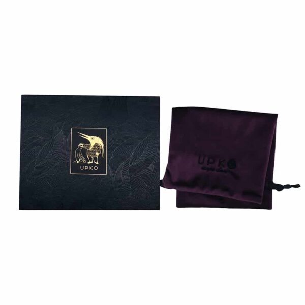 El cinturón bondage UPKO se presenta en una bolsa de terciopelo morado con las letras UPKO en negro. La caja tiene un fondo negro y plateado con motivos de follaje y un cuadrado central con el logotipo de UPKO en dorado.