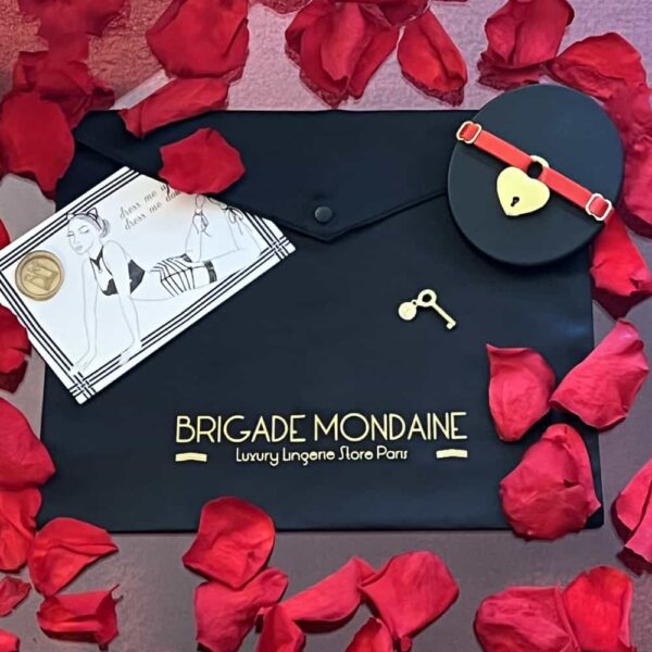 black packaging of the brigade mondaine. On the fabric is inscribed the brigade mondaine gold color. Accompanied by a card, a red choker and rose petals.