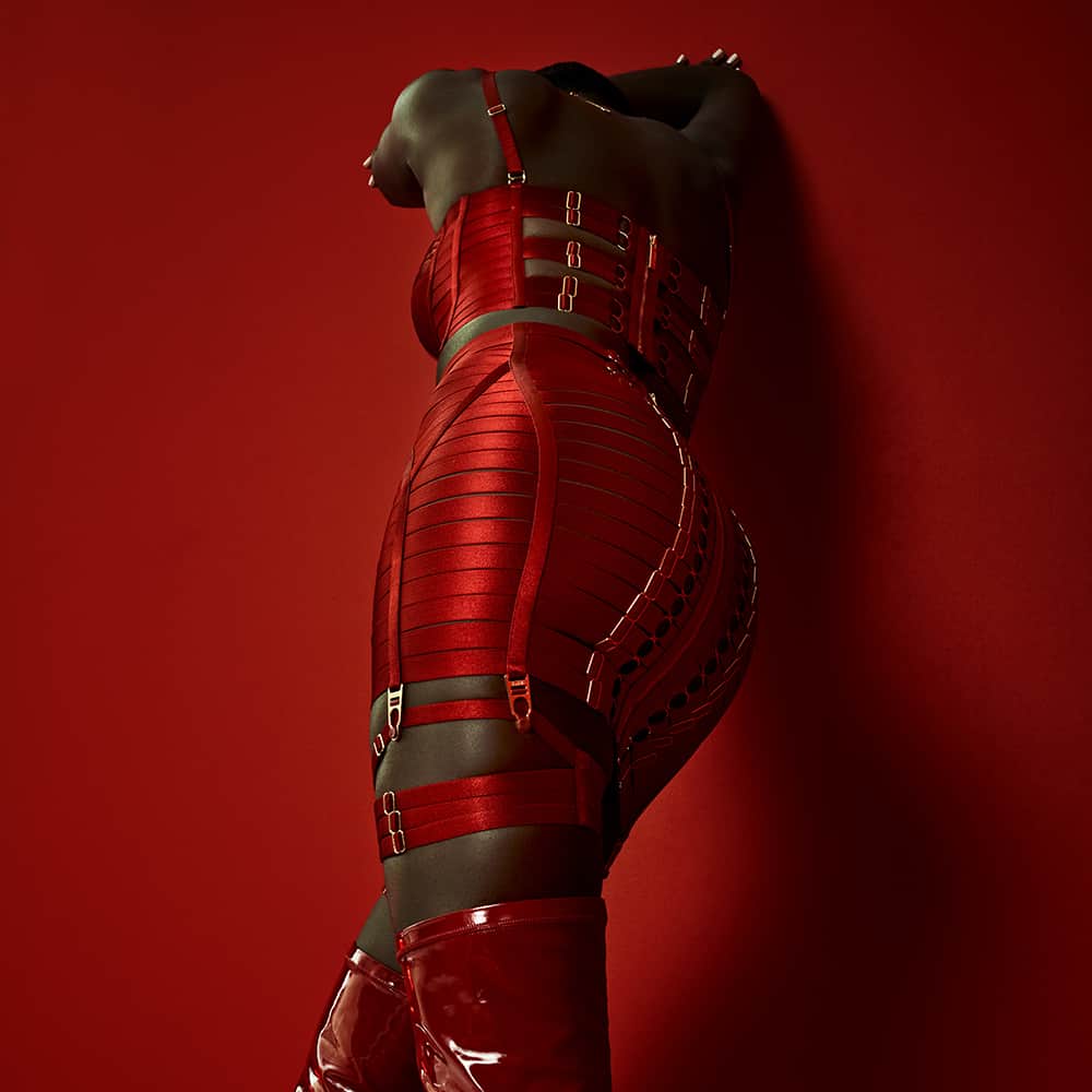 La modelo lleva un conjunto Signature Bordelle en rojo. Se compone de un corpiño Minerva Ruby y un Waspie Ruby Clásico Exclusivo, ambos de raso rojo y elástico de rejilla y chapado en oro.
