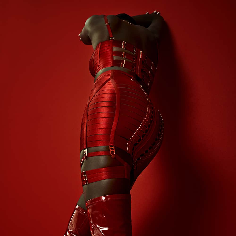 The model is wearing a Signature Bordelle set in red. It is composed of a Minerva Ruby bustier and an Exclusive Classic Waspie Ruby, both made of red satin and fishnet elastic and gold plating.