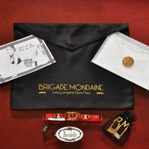 Foto des roten Geschenkpakets Bordelle und Brigade Mondaine. Zu sehen ist eine rote Halskette Bordelle aus Gummiband und einem runden goldenen Detail.