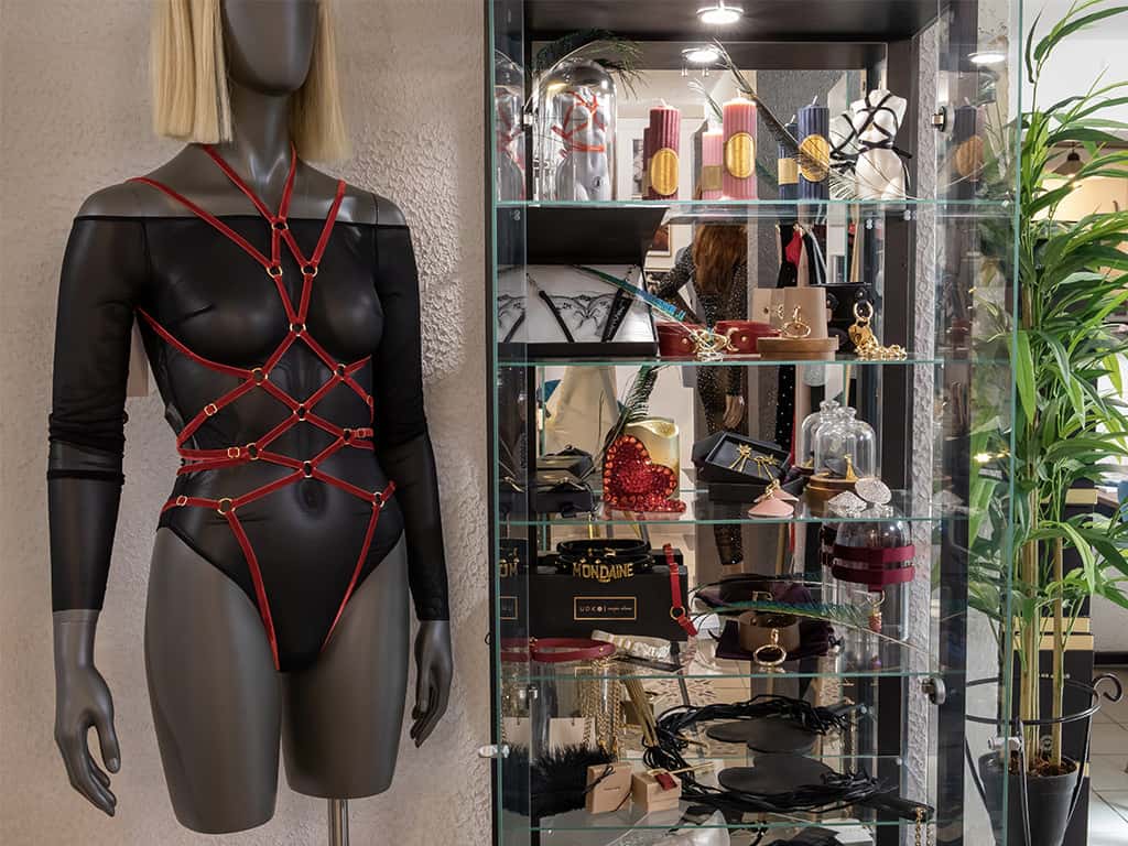 Wir sehen eine Vitrine, die mit verschiedenen BDSM-Accessoires und Kerzen gefüllt ist. Auf der linken Seite des Schaufensters trägt ein Model einen schwarzen Mesh-Body der Marke Zhilyova sowie einen Couture de Nuit-Playsuit aus dünnem, rotem Gummiband.