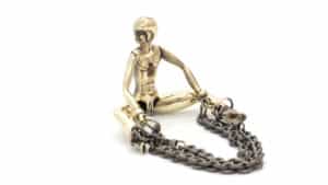 Se trata de un anillo en forma de muñeca de bronce patinado. Mientras gira la cabeza hacia la derecha, abre las piernas y tiene cadenas en los pies. 