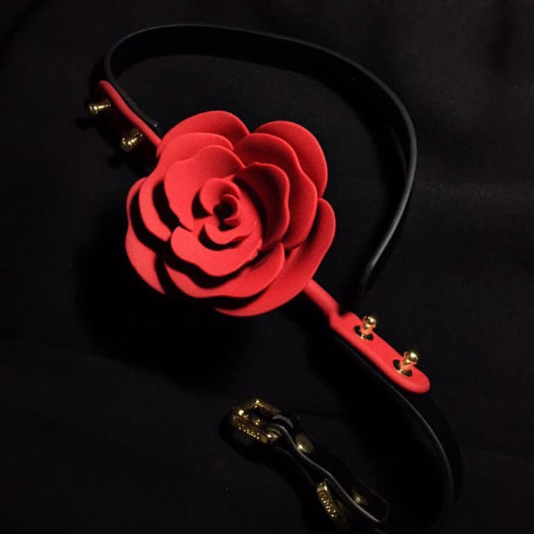 Pink Ball Gag from the brand Upko x Zalo. The product is detached, the back of the product is black and the front is red. The center of the Ball Gag is shaped like a red rose. The whole is placed on a black background.