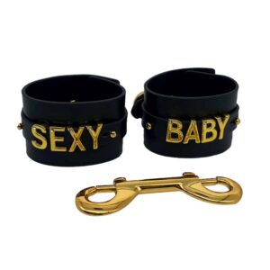 Handcuffs in black Italian leather with hook and letters in 24 karat gold plated and a small stone inlaid on each letter writing the words SEXY BABY from the UPKO X Brigade Mondaine collection available at Brigade Mondaine