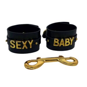 Esposas de suave cuero italiano negro con gancho y letras bañadas en oro de 24 quilates y una pequeña piedra incrustada en cada letra que escribe las palabras SEXY BABY de la colección UPKO X Brigade Mondaine disponible en Brigade Mondaine