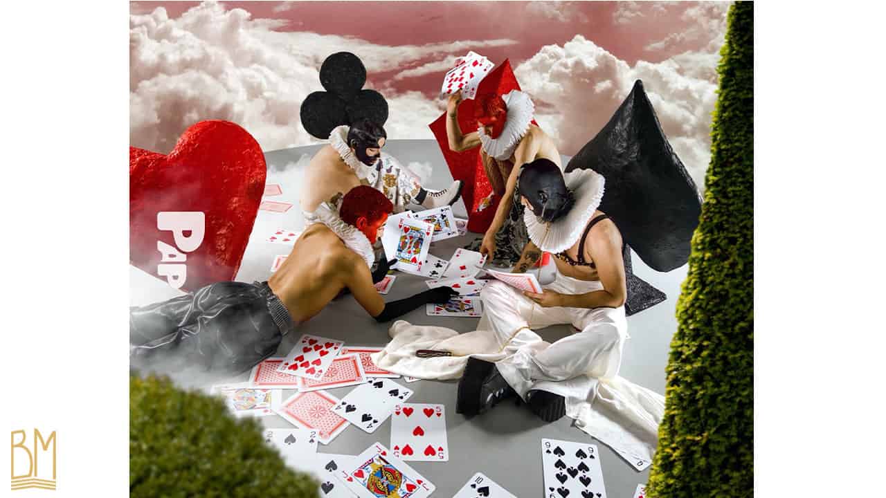 PAP Magazine with 4 men including three men sitting and one lying on the ground. Two of them have the face in black and the other two in red. The man on the right is wearing an Upko leash. They are playing cards. Behind them are the signs for hearts, spades and diamonds. On the floor, there are cards scattered around. Behind them, you can see clouds and the sky.