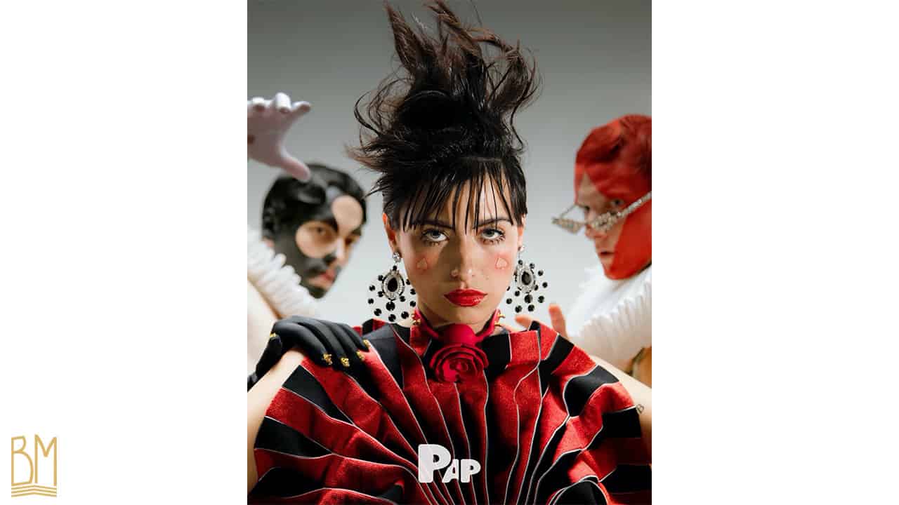 PAP Magazine il s’agit d’une femme portant autour de son cou elle porte le Gag Ball Upko. Sur sa joue, il y a le signe du pique. Derrière elle se trouve deux hommes avec le signe du trèfle et du carreau peints sur leur visage.