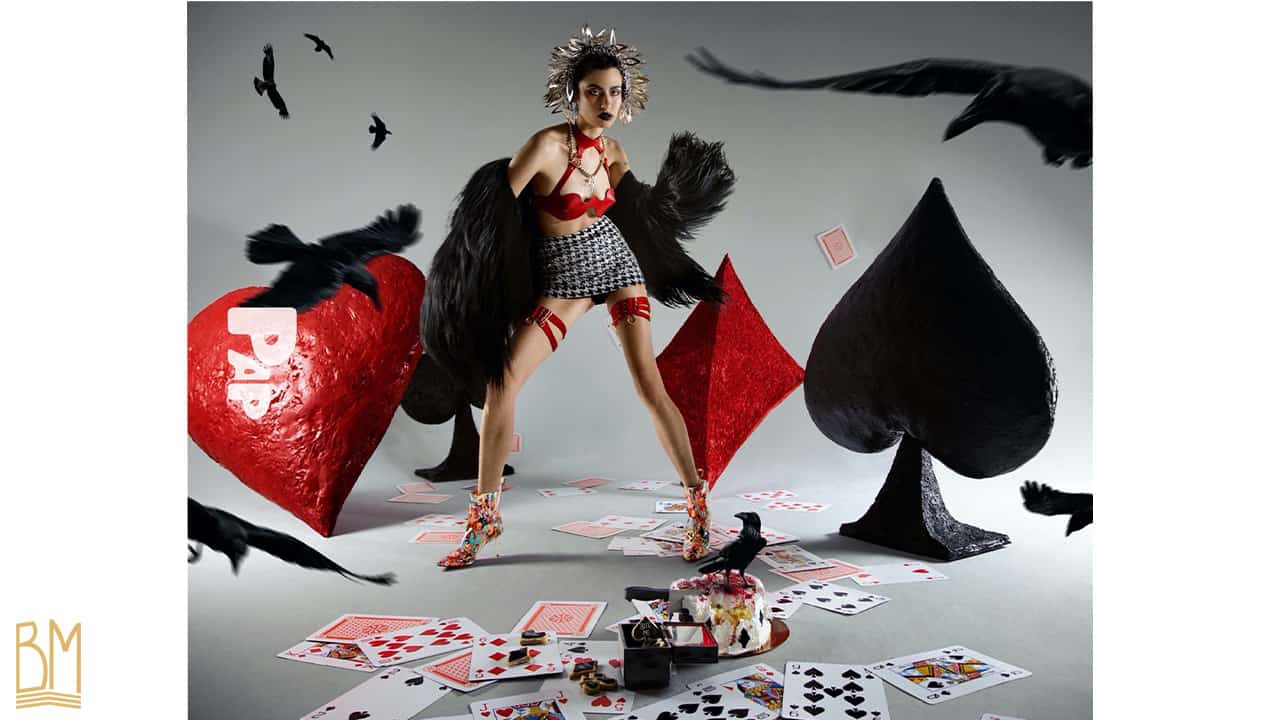 PAP Magazine woman wearing a bra Whiplash Cuir red and red suspenders of the brand ELF ZHOU LONDON. Over it she wears a black jacket with hair. Behind her is the sign of hearts, clubs, spades and diamonds. On the floor are scattered cards and a partially eaten cake with a raven next to it. Other crows are flying around the model.