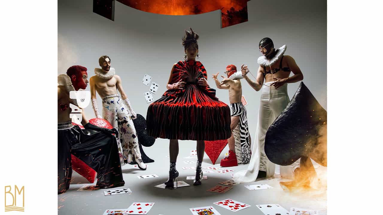 PAP Magazine there are 4 men dressed in black, red and white positioned around a standing woman wearing a red and black dress. The man on the right is wearing an Upko leash. In front of each man there is a red heart, a black clover, a red diamond, a black spade. On the floor are scattered cards.