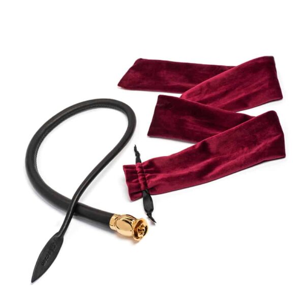 Whip Thorn of the brand UPKO X ZALO gold and black color. The product is a black leather whip with a semi-open rose shaped pommel in gold plated. The end of the whip has a leaf shape with the brands logo embossed. Next to it is the red velvet packaging with black ribbons.