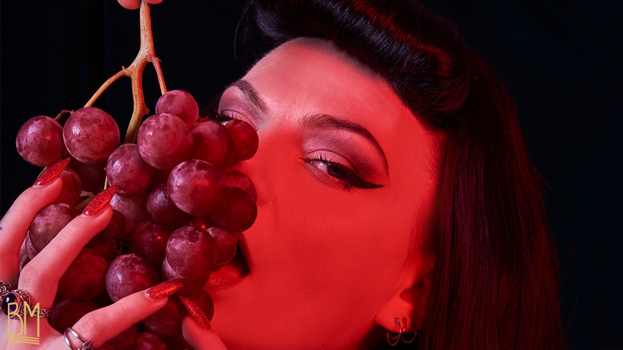 Foto von Julie Von Trash, erstellt von Studio Volua Paris. Sie hält in ihrer rechten Hand eine große Traube roter Weintrauben und schaut in die Kamera. Das Foto zeigt nur ihr Gesicht und betont insbesondere ihren Blick.