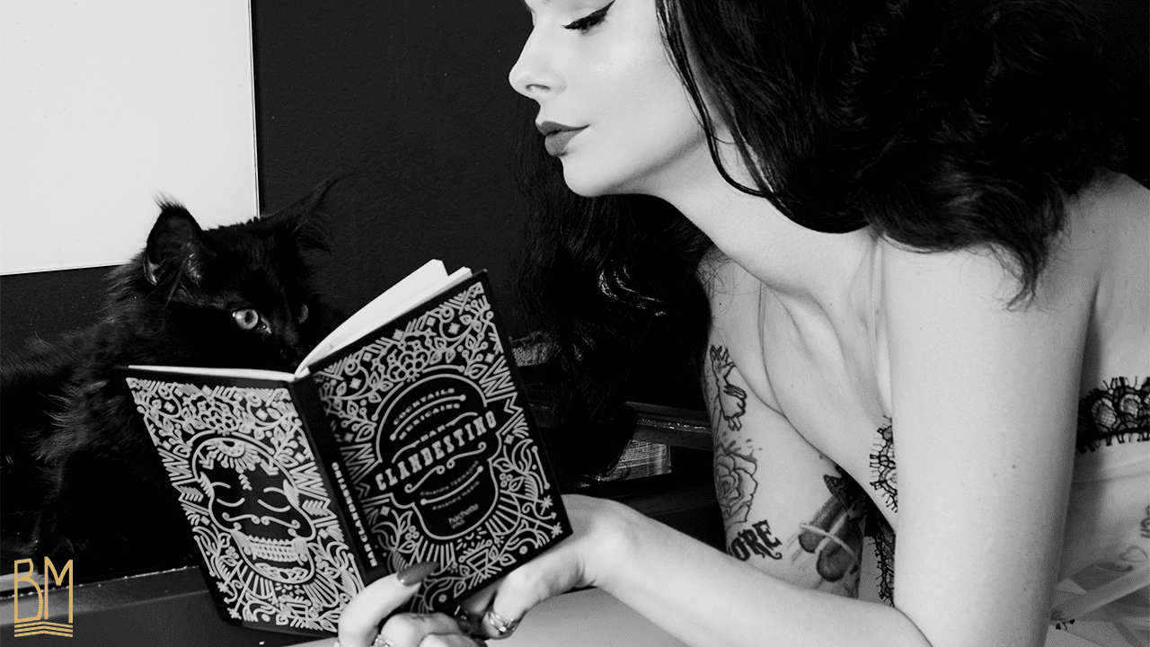 Photo de Julie Von Trash en noir et blanc réalisée par le Studio Volua Paris. Elle porte un kimono de la marque Ludique Lingerie. Elle est couchée et lit un livre. À sa droite, il y a un chat noir.