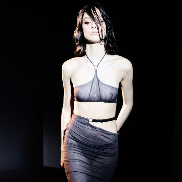 Топ Khan Collection Caravasar от Gonzales Affaires. Этот топ представляет собой бандо с оголенными плечами, которое застегивается на затылке с помощью тонких простых серых бретелей. Серое кольцо соединяет ремешок и лямки на шее. Нижний шов бандо имеет вырез. Спина полностью оголена. Изделие застегивается двумя маленькими крючками на талии. Два больших тюлевых чехла поддерживают бюст по бокам.