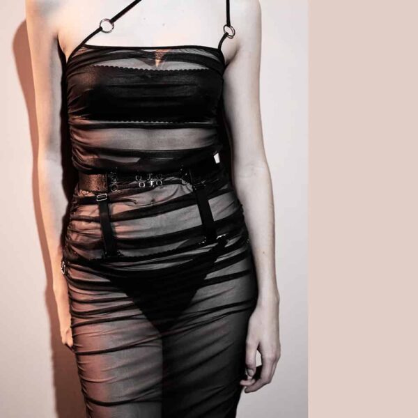 Ensemble Top et jupe Les Indolentes Collection Caravasar de la marque Gonzales Affaires. Cette jupe s’arrête en dessous des genoux. Le produit est confectionné à partir d’une partie en tulle transparent gris des genoux jusqu’au nombril. Une couture est située sur le côté de la robe afin d’obtenir ou non un rendu plissée de la jupe en saisissant l’anneau au bout de la jupe. Ensuite une ceinture est placée au niveau des hanches avec trois élastiques fins ornés d’anneaux argenté au centre de la ceinture. Les deux parties sont reliées par deux bandes élastiqués qui sont amovibles par le biais de deux crochets. Ce top est en tulle transparent gris. Un effet frappé est présent au niveau du ventre. Une couture sur le coté droit du top permet d’obtenir cet effet de manière encore plus prononcé lorsque l’on tire sur l’anneau à l’extrémité de cette couture. Les bretelles se rejoignent toutes les deux sur l’épaule et donnent de ce fait un aspect asymétrique. Deux anneaux argentés agrémentent ces bretelles. À l’arrière, le haut du dos est nu et les deux bretelles se rejoignent en une seule par le biais d’un anneau puis d’une bande élastiquée réglable qui est cousue sur le top. Le top et la jupe se juxtaposent parfaitement et forment une robe en tulle d’une grande élégance.