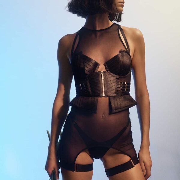 Black ensemble from the Kora collection by Bordelle, the pieces in the Kora collection are handcrafted from removable elements to be worn in a multitude of ways. The Kora basque is the signature piece of this collection, designed as an outerwear centerpiece with structured and molded cups. All the elastic straps are adjustable. The model wears the basque over the black garter dress, in fine fishnet with a fully open back to the kidneys.
