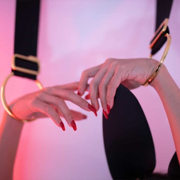 Mains de femme vernis, retenues au-dessus de sa tête par des larges sangles noires avec des attaches dorées aux extrémités. Les poignets sont à l'intérieur d'anneaux dorés.