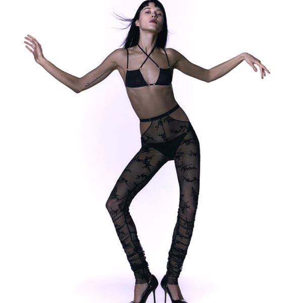 Iggy Collection Caravasar by Gonzalez. Sheer black pants with lace pattern and silver elastic straps. The bralette is triangle-shaped and black. Two small silver-colored rings are placed at the end of the cups at solar plexus level and hold two small decorative elastic straps. The cups are made of tulle and the straps are thin. The bralette fastens at the back with a simple hook.