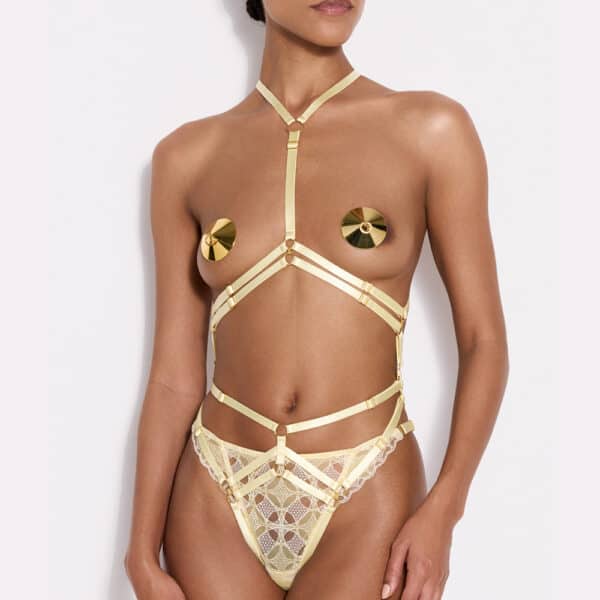 a front-facing woman with dark-colored skin wearing a harness with several interconnected straps and a yellow lace thingy and gold-colored nipples