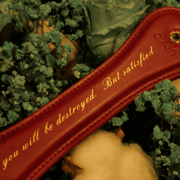 Fotografía de la Upko Paleta de cuero rojo con letras doradas que rezan "Serás destruido. pero satisfecho". La paleta roja está engastada en un ramo de flores azules.
