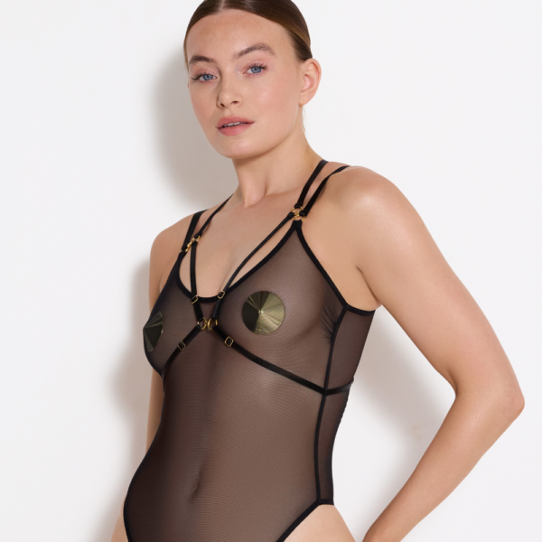 Photograph in which a woman in front of a white wall wears a transparent black bodysuit with harness, straps and gold detailing in addition to nipples to hide her nipples.