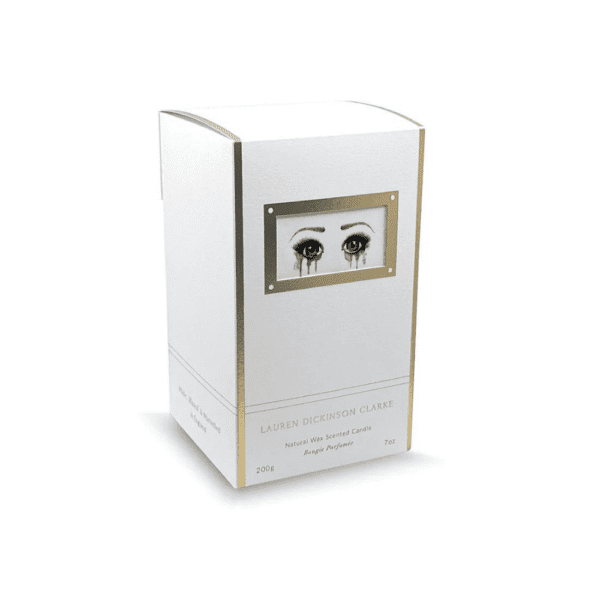 The candle's packaging is white with gold accents, and features a design of sad eyes.