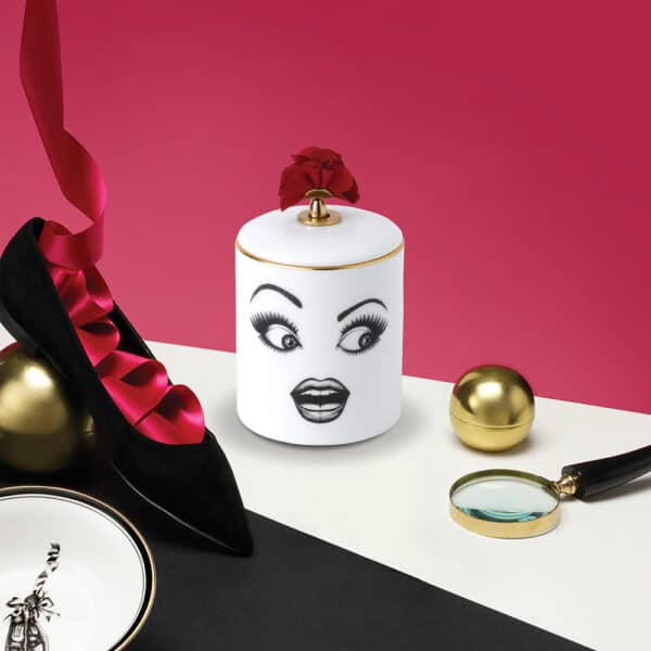Muse Parfumée candle A surprised face, delicately drawn with felt on white porcelain adorned with gold and red details, placed on a white table set against a pink wall featuring a ballerina and ribbon.