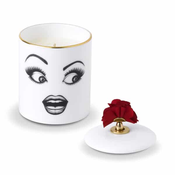 Muse Parfumée candle A surprised face, delicately drawn with felt on white porcelain adorned with gold and red details, placed on a white table set against a pink wall featuring a ballerina and ribbon.