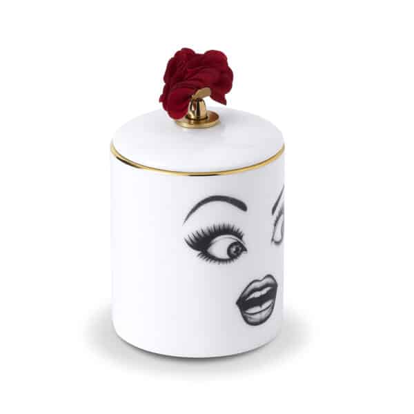 Muse Parfumée candle A surprised face, delicately drawn with felt on white porcelain adorned with gold and red details, placed on a white table set against a pink wall featuring a ballerina and ribbon.