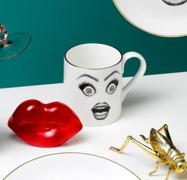 Eine weiße Tasse aus chinesischem Porzellan zeigt eine schwarze Filzzeichnung, die ein überraschtes Gesicht mit sehr schockierten Augen und einem offenen Mund illustriert, das mit goldenen Details akzentuiert ist.