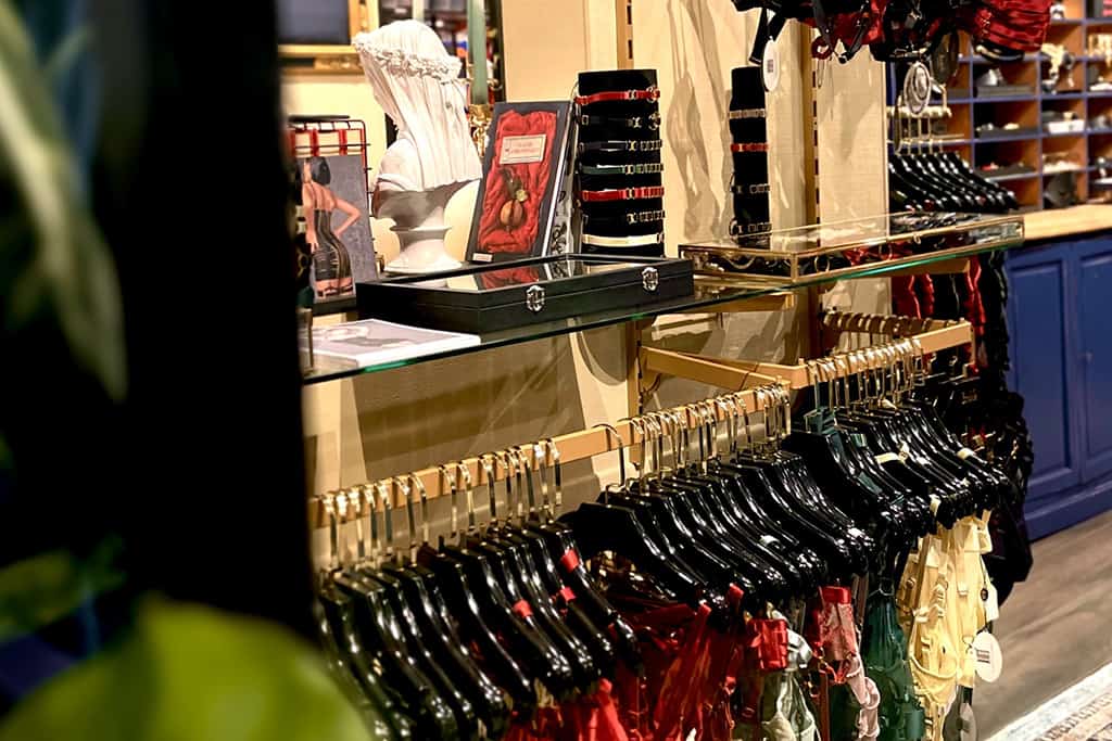 A photograph taken from a distance reveals a clothing rack on which several colorful pieces are carefully arranged in shades of yellow, green and red, suspended from lacquered black hangers. Above this rack, on glass shelves, stand out two black velvet racks, showcasing necklaces, and a transparent box with gold edging.