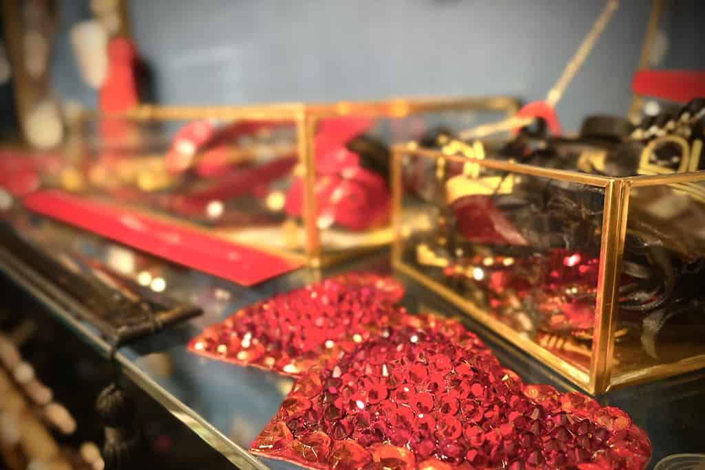 A close-up photograph shows two transparent boxes adorned with gold details. In the foreground are two red hearts encrusted with rhinestones. Inside the boxes, red objects and black pouches can be seen.