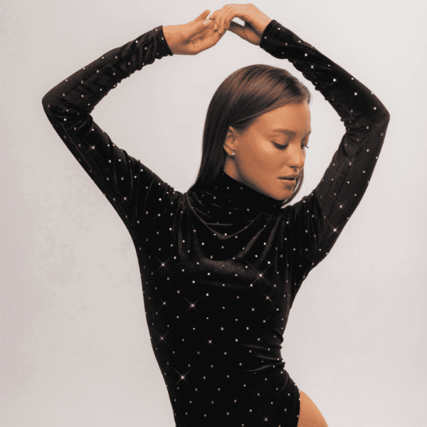 Una mujer posa con elegancia frente a una pared blanca, vestida con un body de terciopelo negro de cuello alto. Esta refinada prenda se ve realzada por la meticulosa incrustación de cristales de Swarovski, que añaden un toque de brillo y resplandor a su atuendo. La combinación de terciopelo oscuro y cristales brillantes crea un cautivador contraste visual, subrayando la sofisticación y la atención al detalle en sus elecciones de vestuario. Esta combinación única de texturas y elementos decorativos confiere al conjunto un aire chic y moderno, captando la atención y reflejando el refinado gusto de la mujer por la moda.