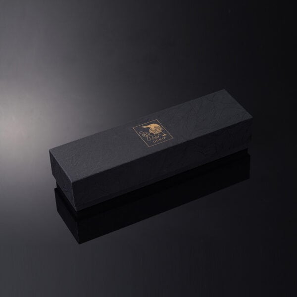 El envase negro y dorado de UPKO está colocado en diagonal sobre un fondo negro brillante que capta y refleja la luz blanca.